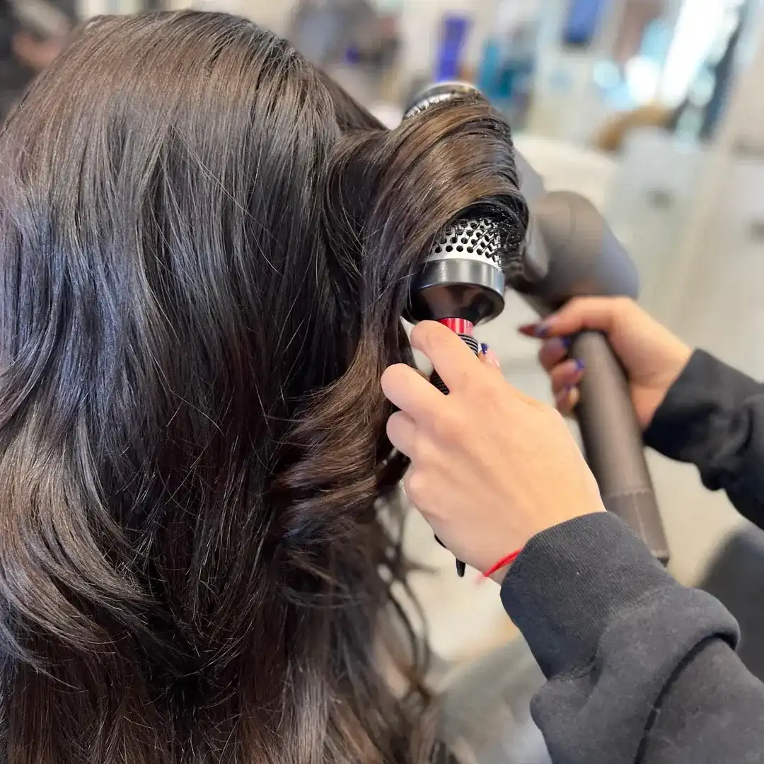Hair stylist curling long, dark hair with a dryer and round brush in a salon setting.