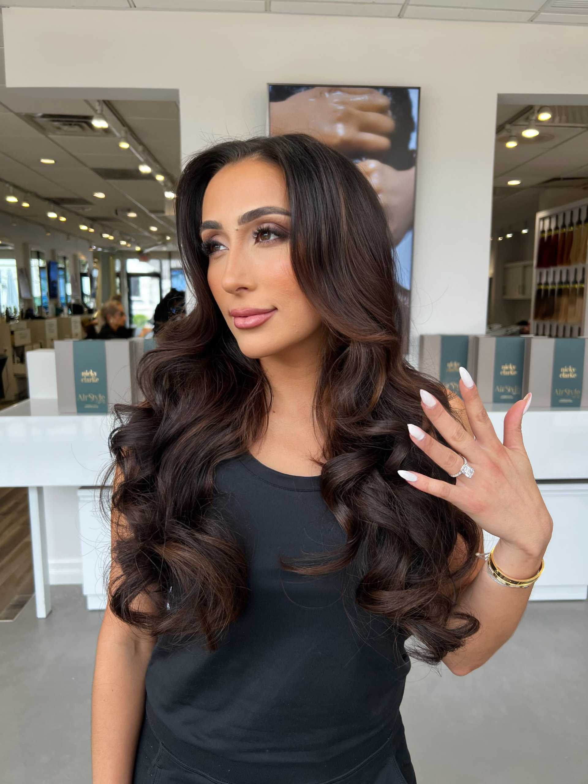 Woman with long curly brown hair posing in a modern salon, showing her ring and nails.
